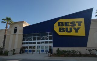Best Buy store exterior at Gulf Coast Town Center with angled blue facade, yellow tag sign, glass entrance and palm trees