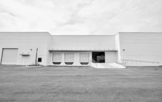 Completed white warehouse with three loading dock doors, open bay, concrete ramp with railings and a large paved lot