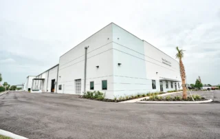 Completed white Balentine Builders manufacturing facility with "Solid Surface Tops" sign, roll-up doors, palm tree and new