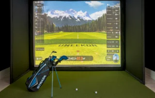 Indoor golf simulator bay at The Mark with Uneekor driving range on screen, turf mat, golf balls and a Strata golf bag