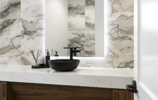 Office restroom with marble-look tile wall, backlit mirror, black vessel sink on white stone counter, and wood vanity